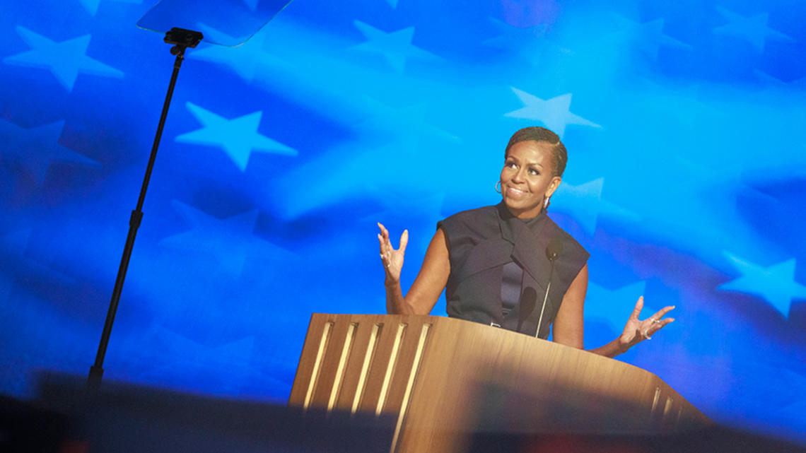 AITN102224_MichelleObama_0.jpg Michelle Obama ’85 speaks at a podium in front of a blue starred background; a teleprompter is in the foreground.