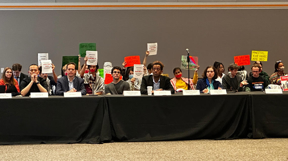 Pro-Palestinian students sit behind members of the CPCU's table holding signs.