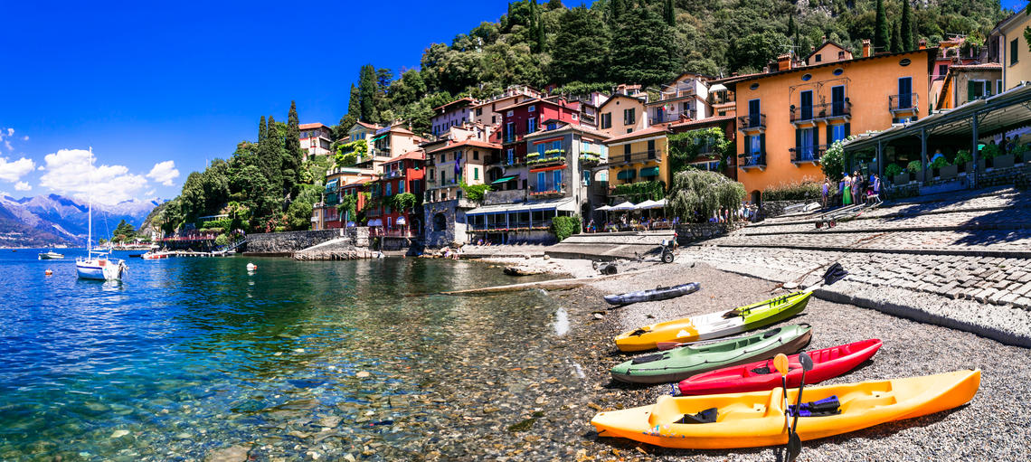 The edge of Lake Como in Italy, with clear water showing pebbles at the bottom, colorful plastic canoes lined up in the foreground, and charming brightly colored Italian homes in the background.