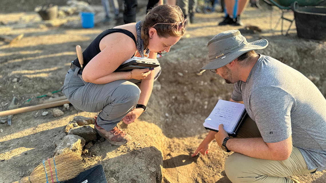 Two people at an archaeological dig examine something in the dirt.