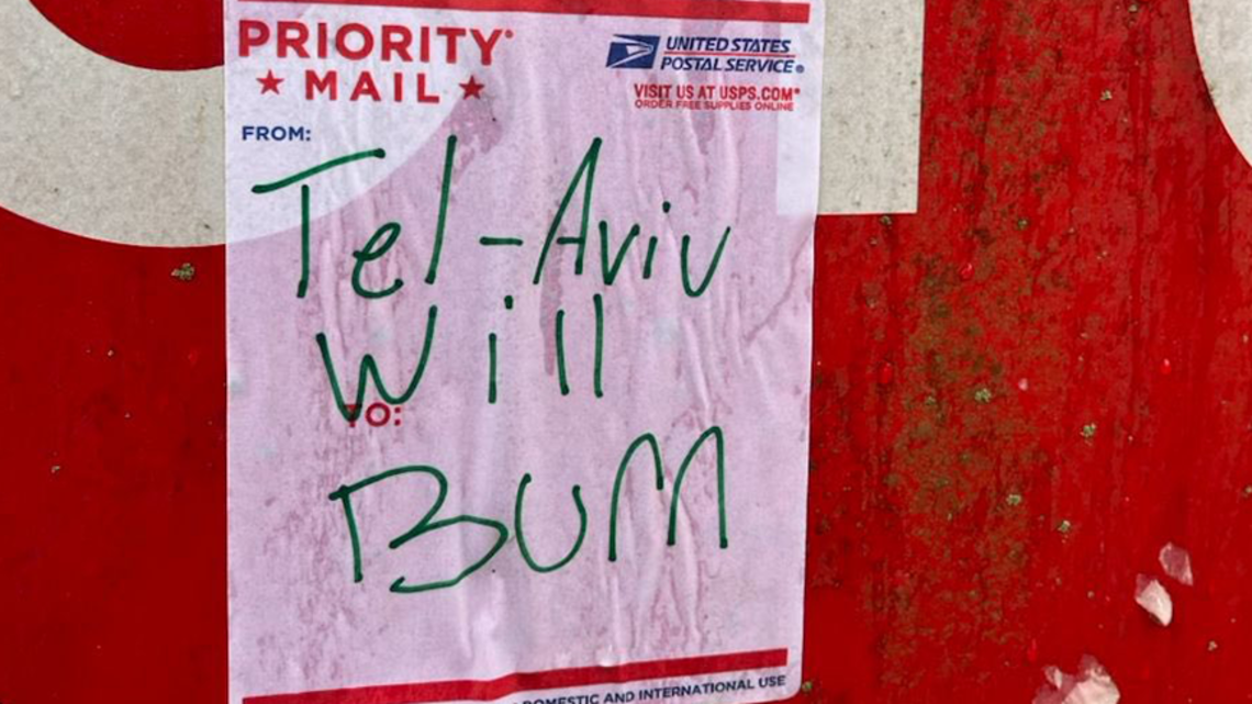 A priority mail sticker from the United States Postal Service stuck to a stop sign. Handwriting in green marker on the sticker says "Tel-Aviv Will Burn"