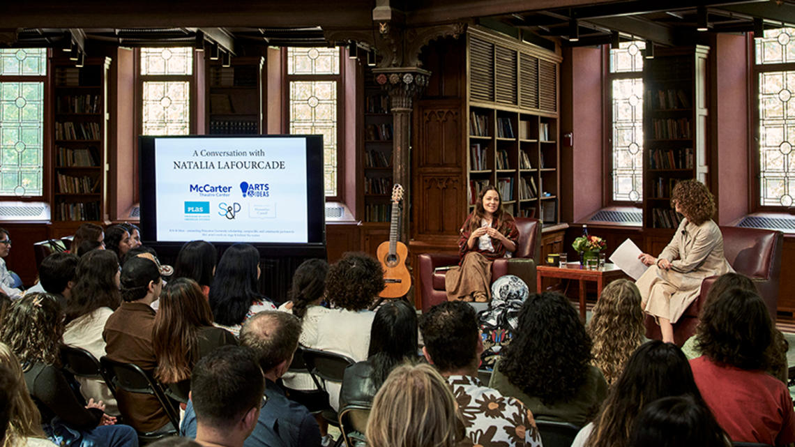 Two women and a guitar next to a screen, speaking to a crowd.