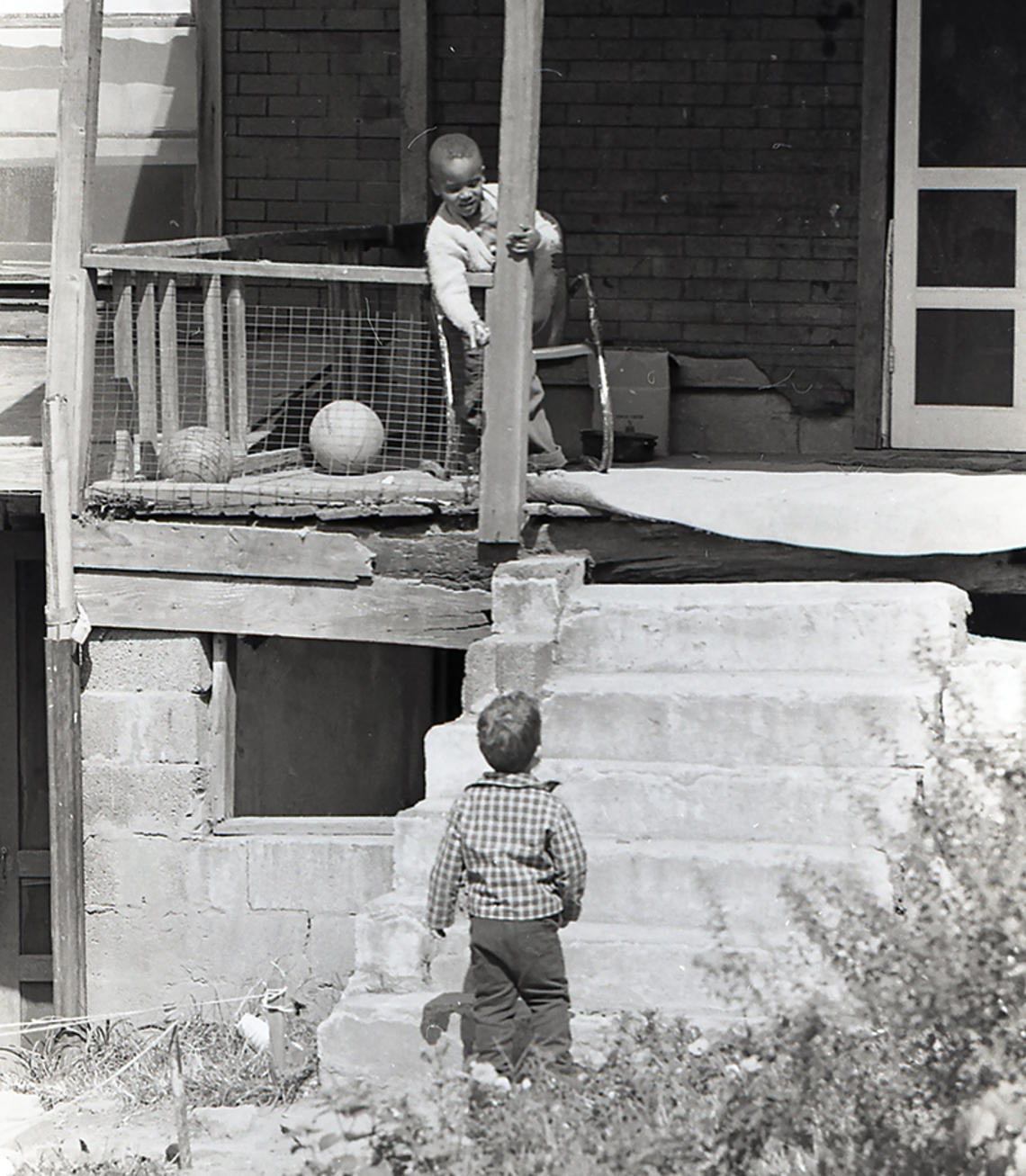 In this black-and-white photo from the 1960s, a boy stands on the porch of a home in rough shape, talking to another boy on the ground.