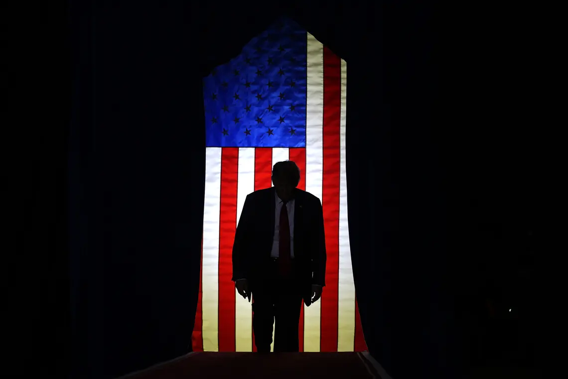 TrumpInFrontofFlag.jpeg Trump standing in front of a lit U.S. flag surrounded by darkeness