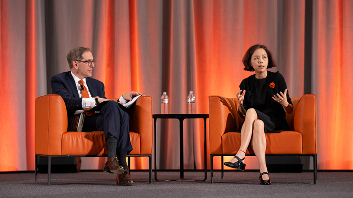 Two people sit on orange chairs on a stage.