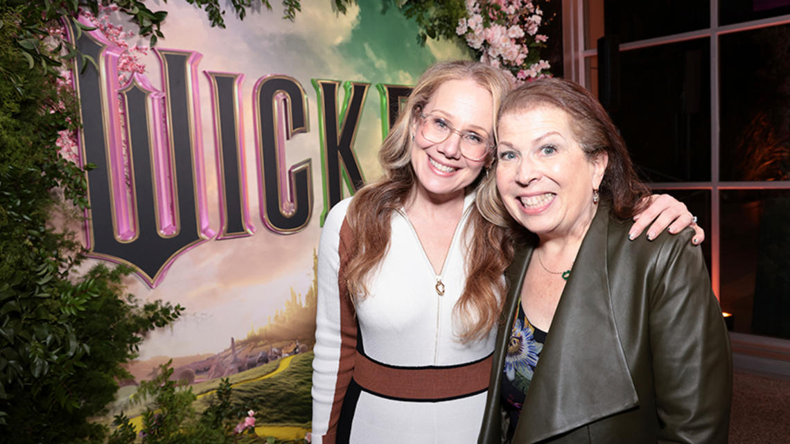 Two women smile for the camera in front of a poster for "Wicked" the movie.