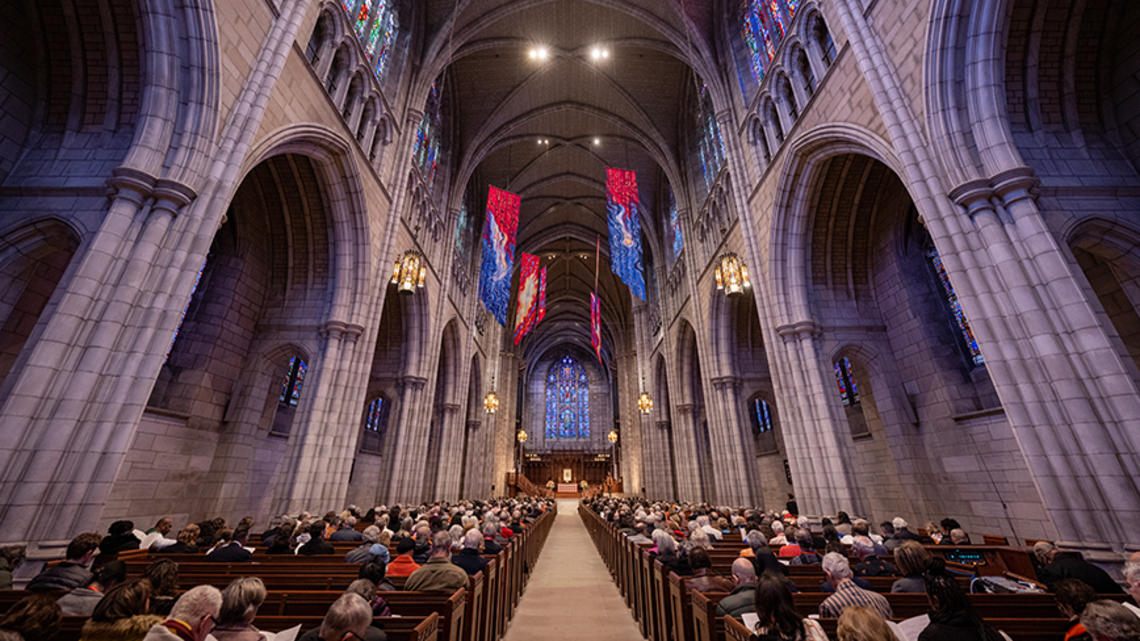 View down the aisle of the Gothic University Chapel, flanked by stone arches and full of people in the pews.