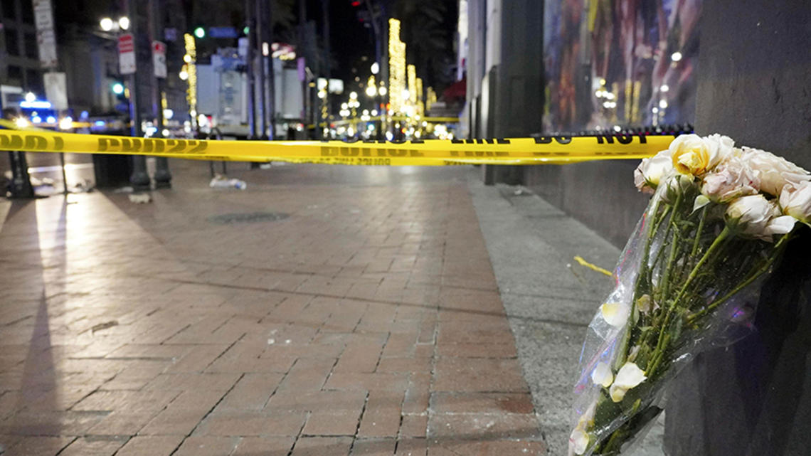 TigerBech_new orleans truck attack.jpg At night, a bouquet of flowers leans up against a building next to a brick sidewalk blocked off with caution tape.