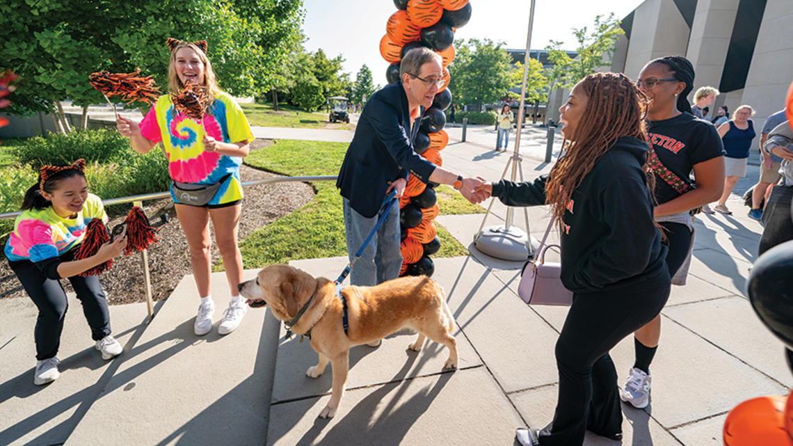 President Eisgruber shakes a woman's hand in front of an orange-and-black balloon arch; his other hand holds the leash of a yellow labrador.