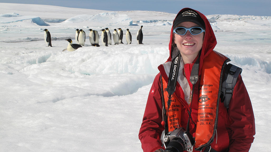 TOW_HeatherLynch_with emperors Credit Evan Grant (1).jpg A woman in a red jacket and sunglasses holds a camera in an ice-covered place, while about a dozen emperor penguins stand in the background.
