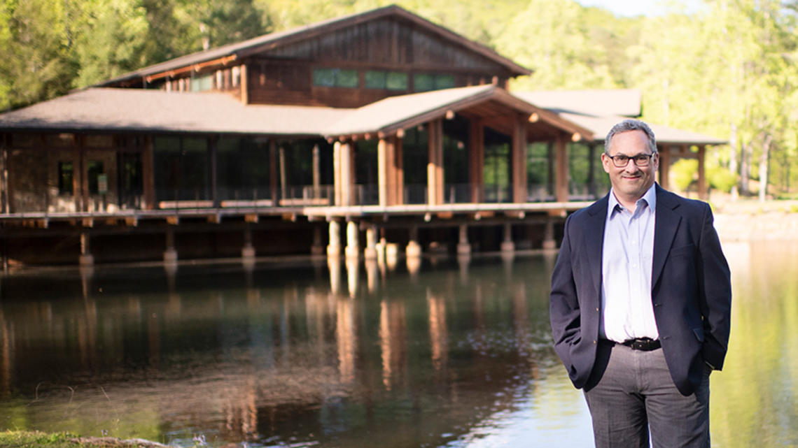 Jason Posnock ’94 stands in front of a pond and a rustic-looking building at the Brevard Music Center.