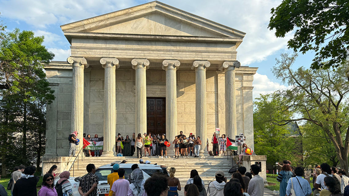 The front of Clio Hall, a classical-Greek-style columned building, with protesters standing on the steps. 