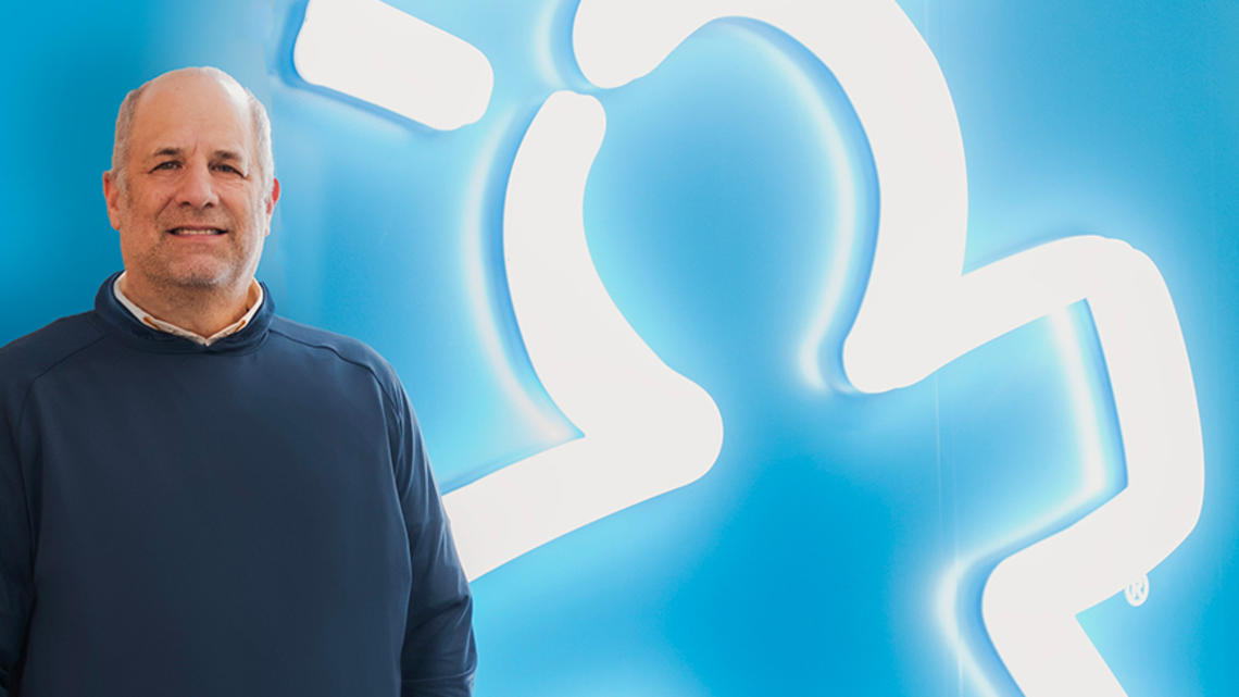 Rick Woldenberg ’81 against a blue background where his company's logo — a person with lines like a lighbulb overhead — is glowing in white neon bulbs.