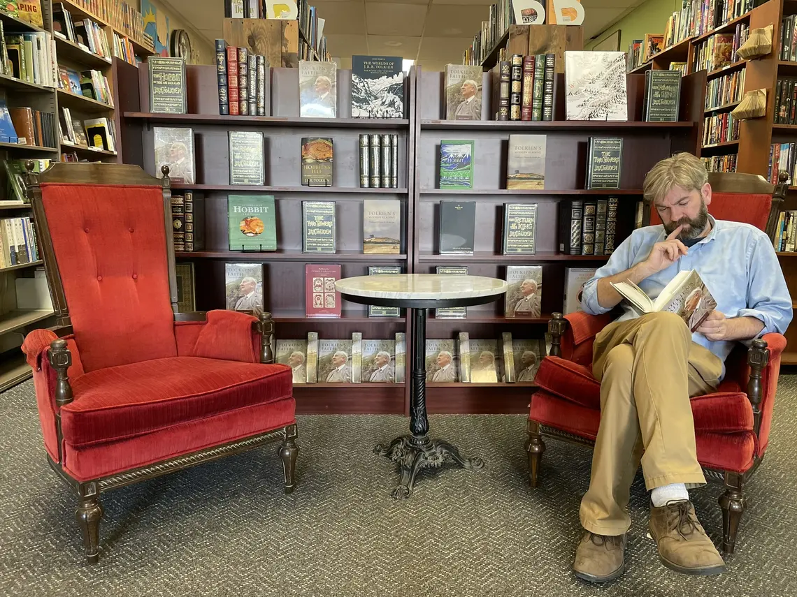 John Kuhner sits at one of two red chairs and reads a book in front of a bookcase in his bookstore.