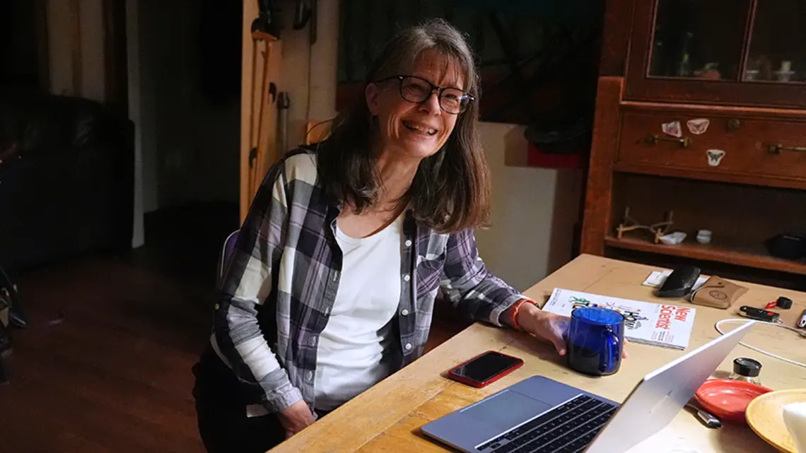 A woman sits at a kitchen table with an open laptop computer.