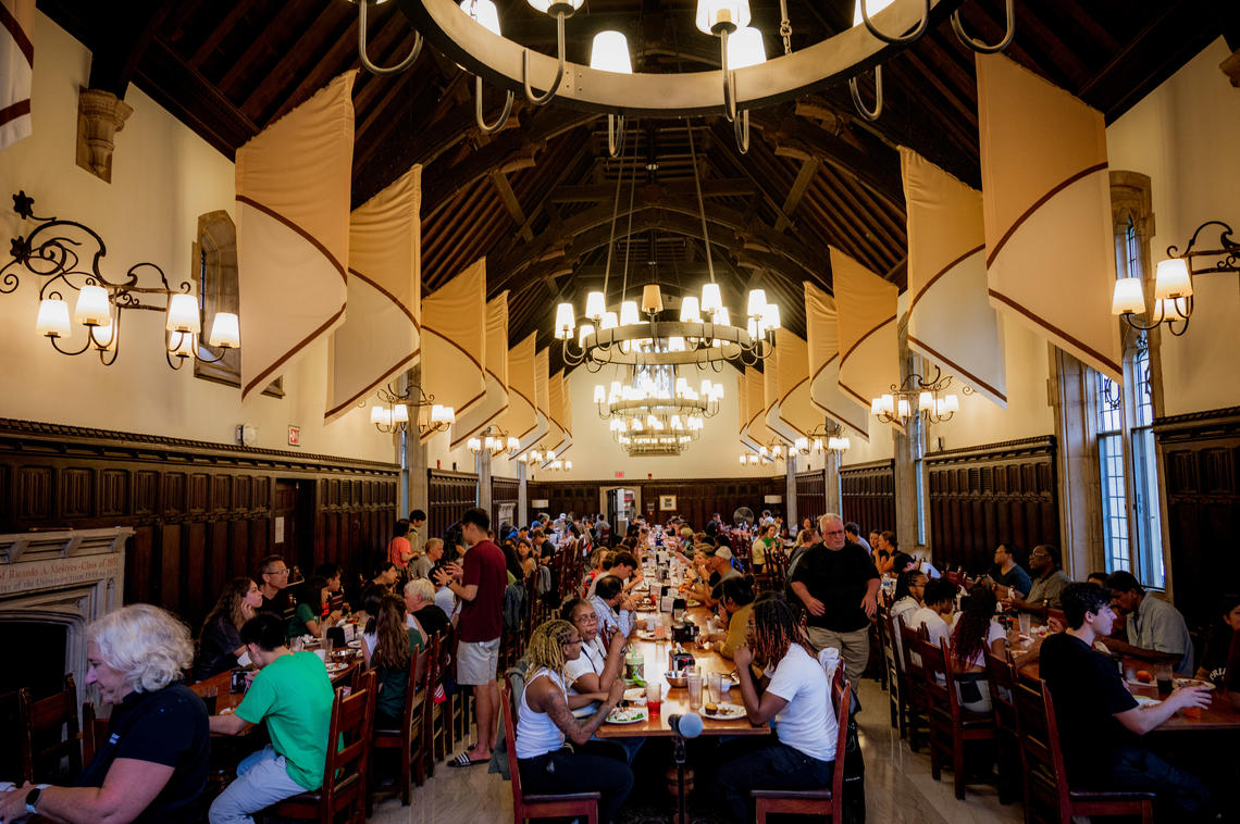 Students sit at long tables arranged in three rows in a dining hall with a high wood ceiling.
