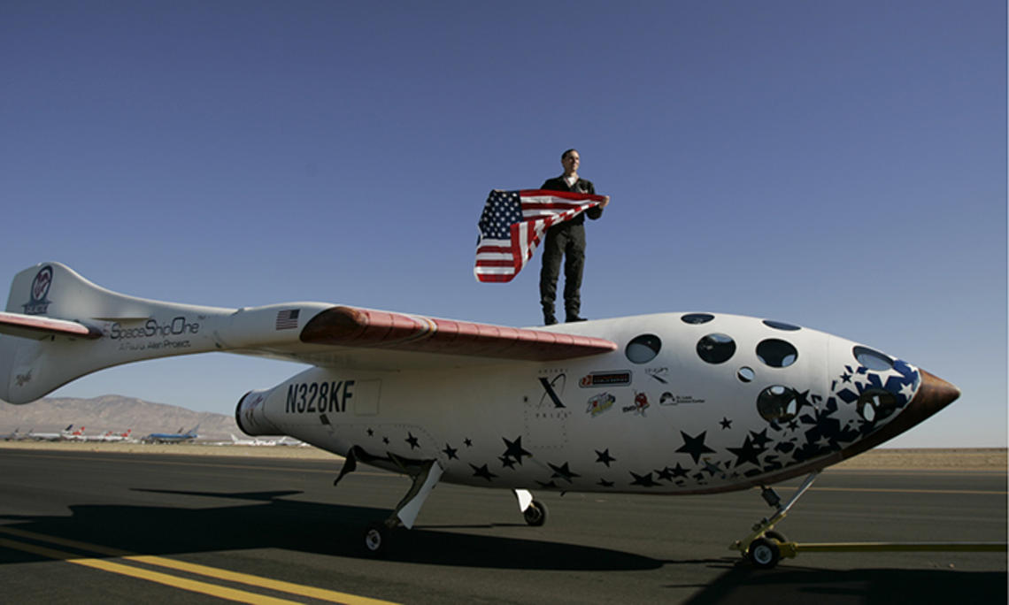 Brian Binnie *78 stands on what looks like a small airplane, holding an American flag.
