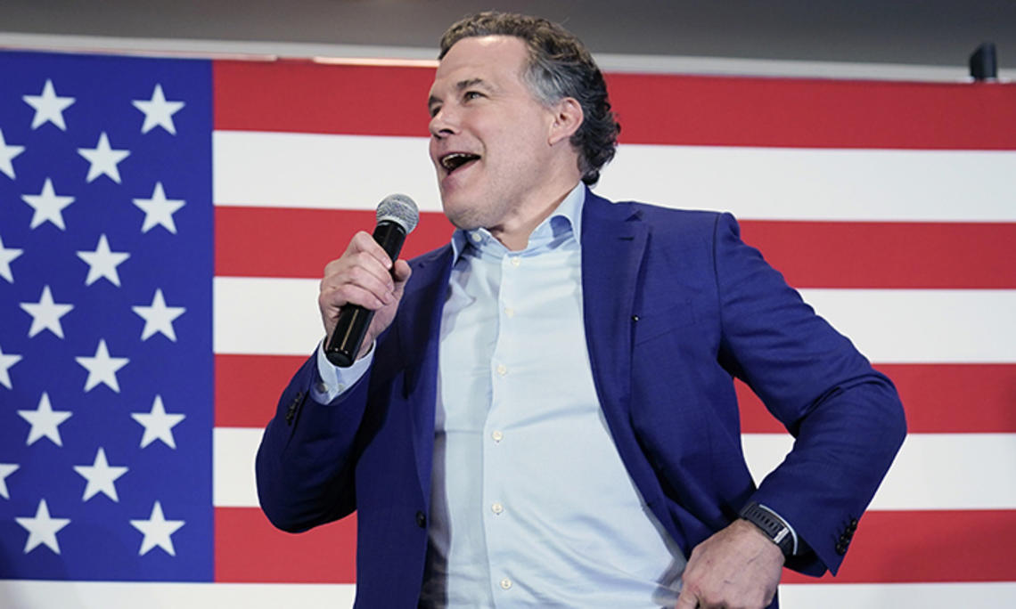 This is a photo of David McCormick speaking into a microphone, with a large American flag in the background.