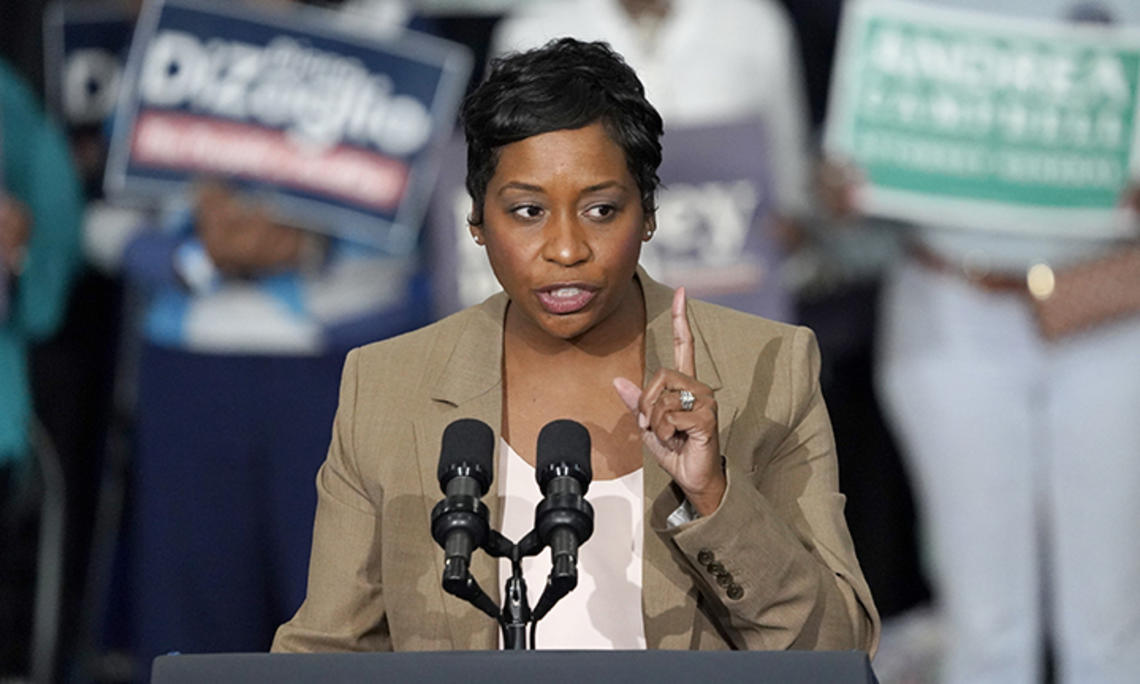 Andrea Campbell speaks at a podium; people hold campaign signs in the background.