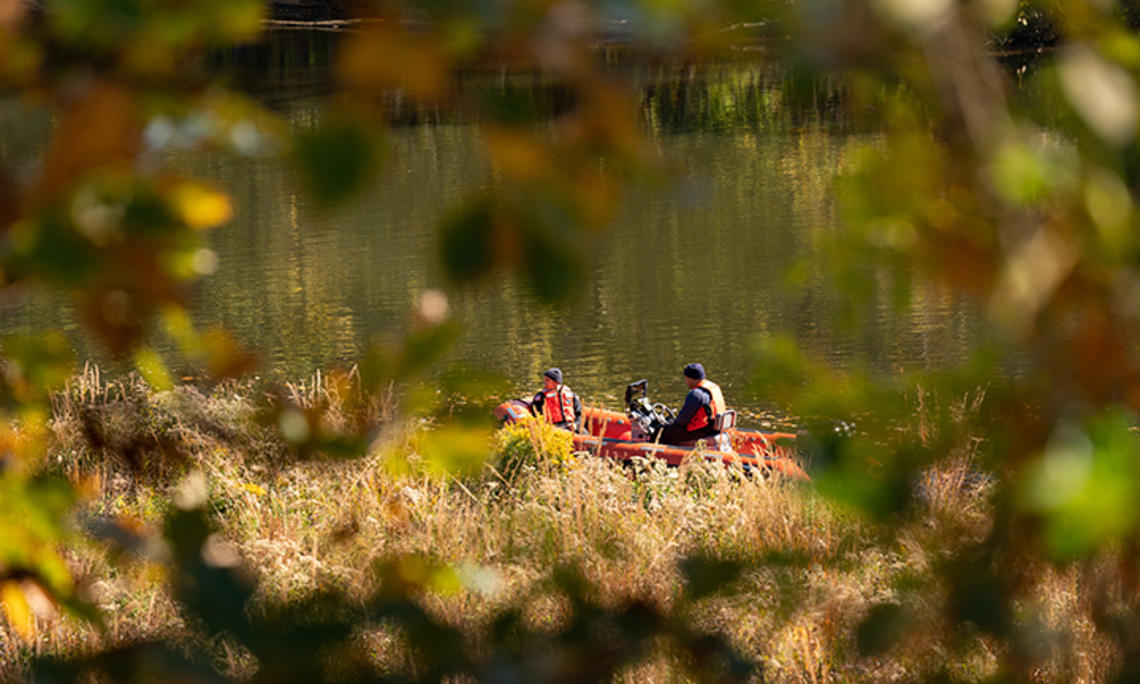 Seen through an opening in the trees, two people in orange vests motor on Lake Carnegie in an orange boat.