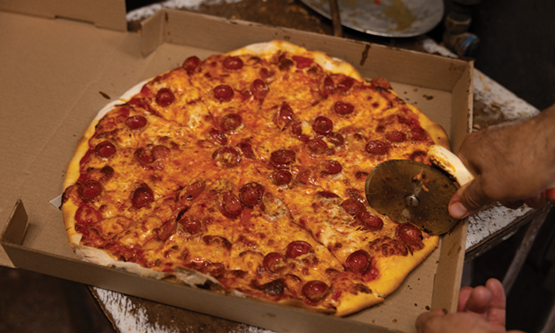 A pizza cutter slices into a pepperoni pizza in a pizza box.