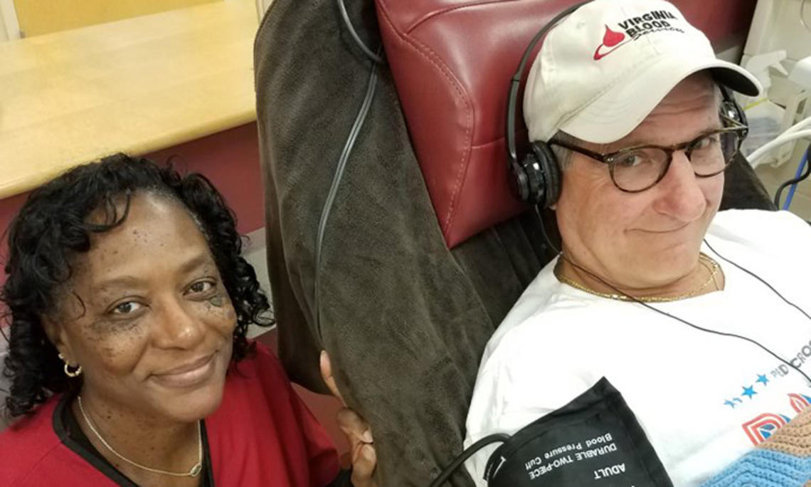Wearing a "Virginia Blood" baseball cap, Doug Schutte ’76 sits in a chair wearing headphones and a blood pressure monitor as a Red Cross worker poses next to him.
