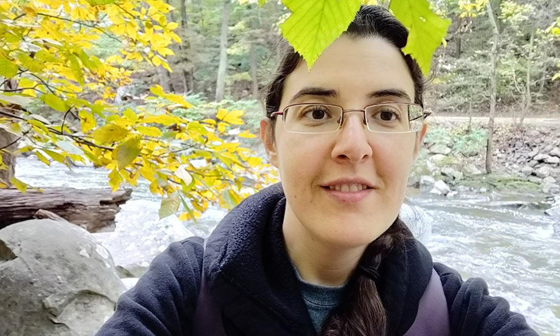 Elizabeth Tsurkov takes a selfie under a tree with a small river behind her.