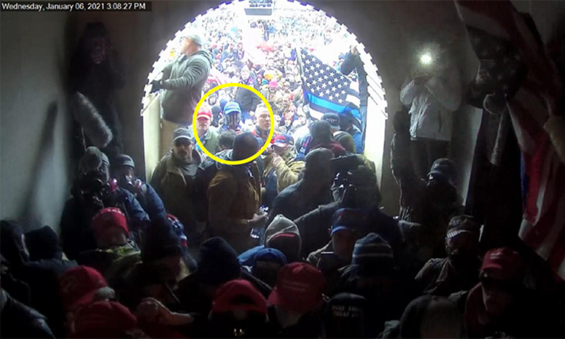 The view out of a crowded Capitol entrance tunnel; a yellow circle highlights Giberson facing the entrance wearing a blue baseball cap and a gaiter over his face.