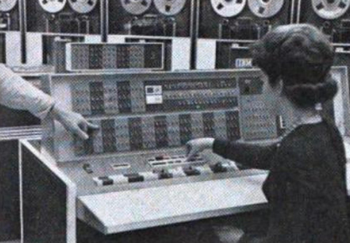 A woman sits at the controls of the room-sized IBM 7090 at Princeton’s Computer Center, manually entering instructions. You can see the data storage tapes at the top of the screen.