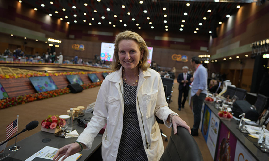 Isobel Coleman ’87 faces the camera and smiles in a room where each seat has a small screen. Blurry in the background are lit-up signs reading G20.