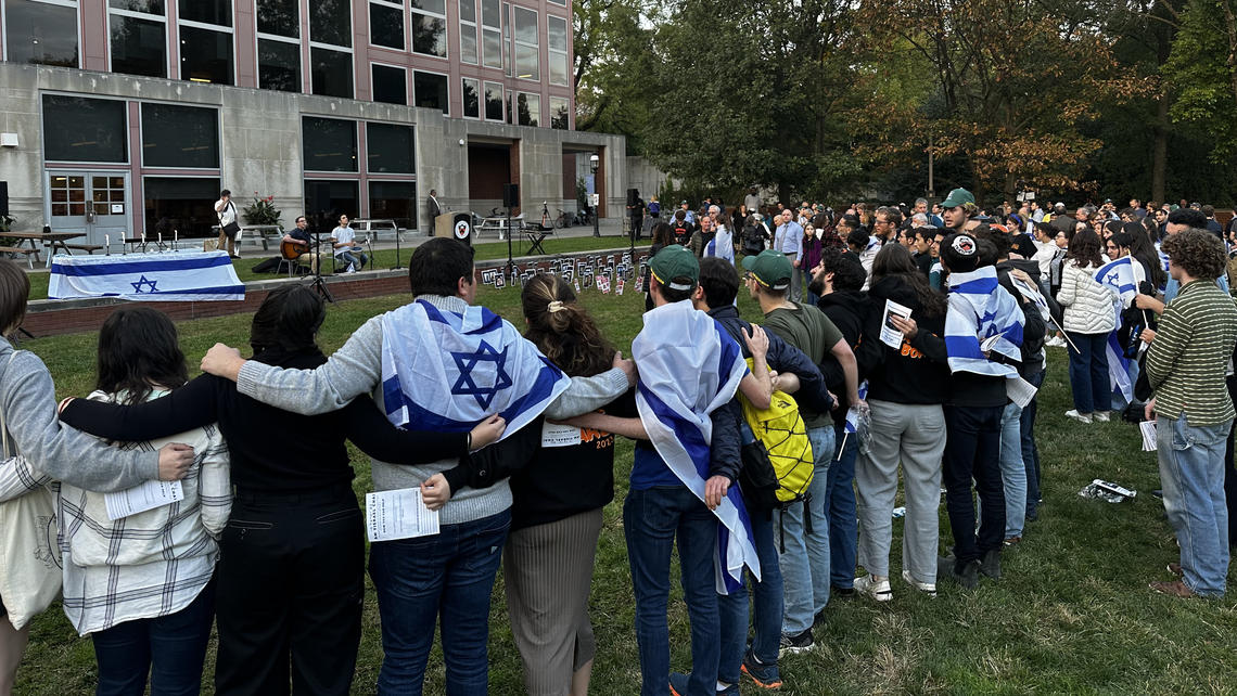 People stand with their arms around each other's shoulders outside a campus building, listening to two students play music. A few have blue-and-white Israel flags wrapped around their shoulders.