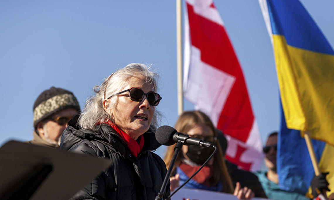 Former U.S. ambassador to Ukraine Marie Yovanovitch ’80 speaks into a microphone during a solidarity rally for Ukraine on Feb. 19, outdoors, wearing sunglasses, with blue sky and the Ukrainian flag in the background.