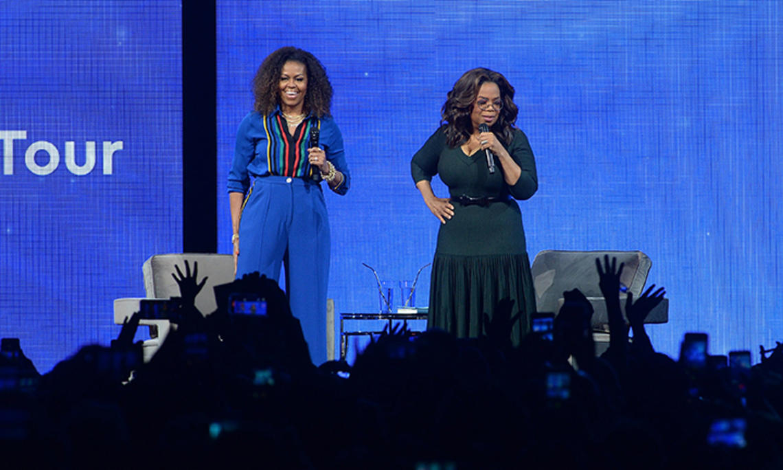 Michelle Obama and Oprah address an audience from a stage.