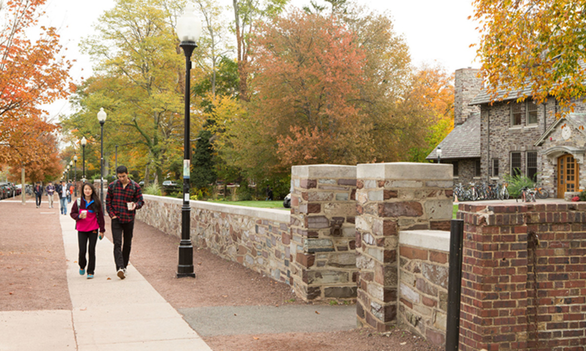 Two students holding coffee cups walk on a sidewalk next to a stone wall. A stone eating club can be seen to the right behind the wall.