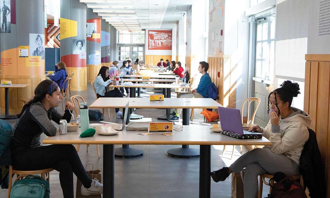This is a photo of students working on laptops and eating in the Frist Campus Center, distanced by sitting longways across from each other, two to a table.