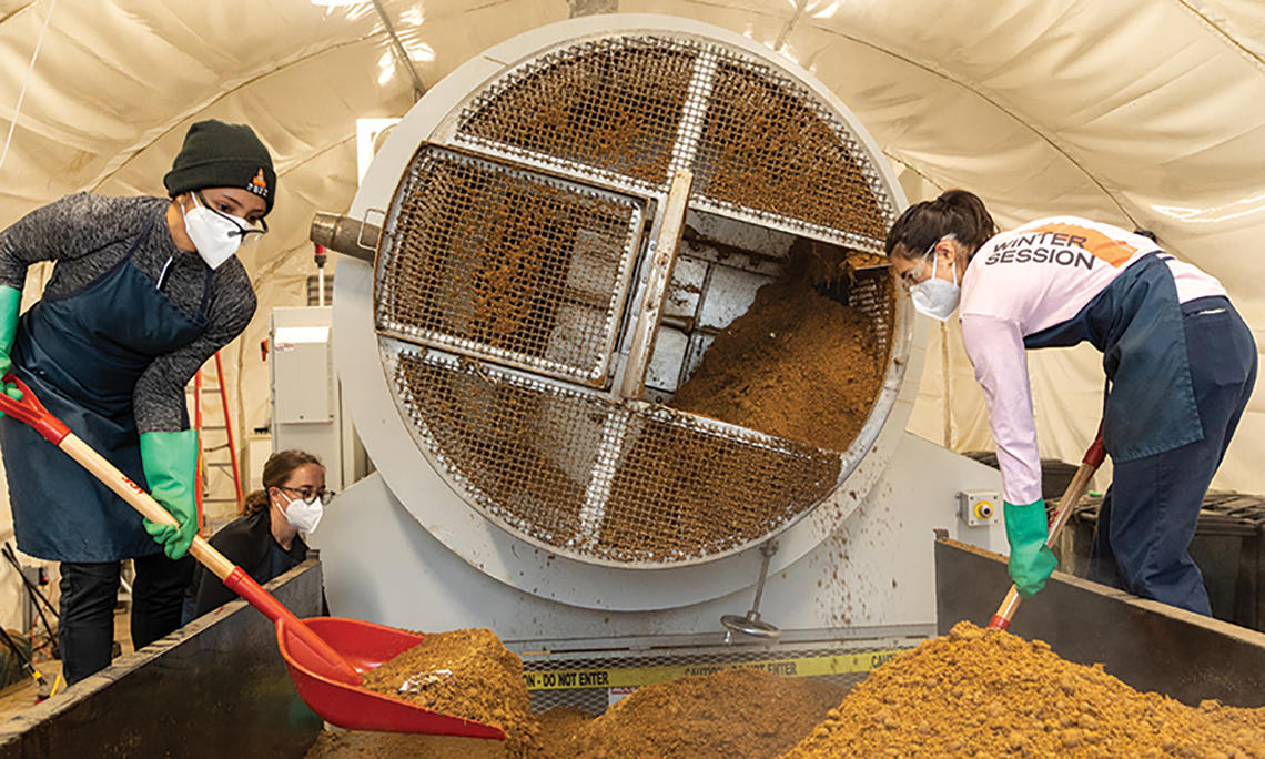 Students shovel compost that looks like dirt next to a cylindrical machine.
