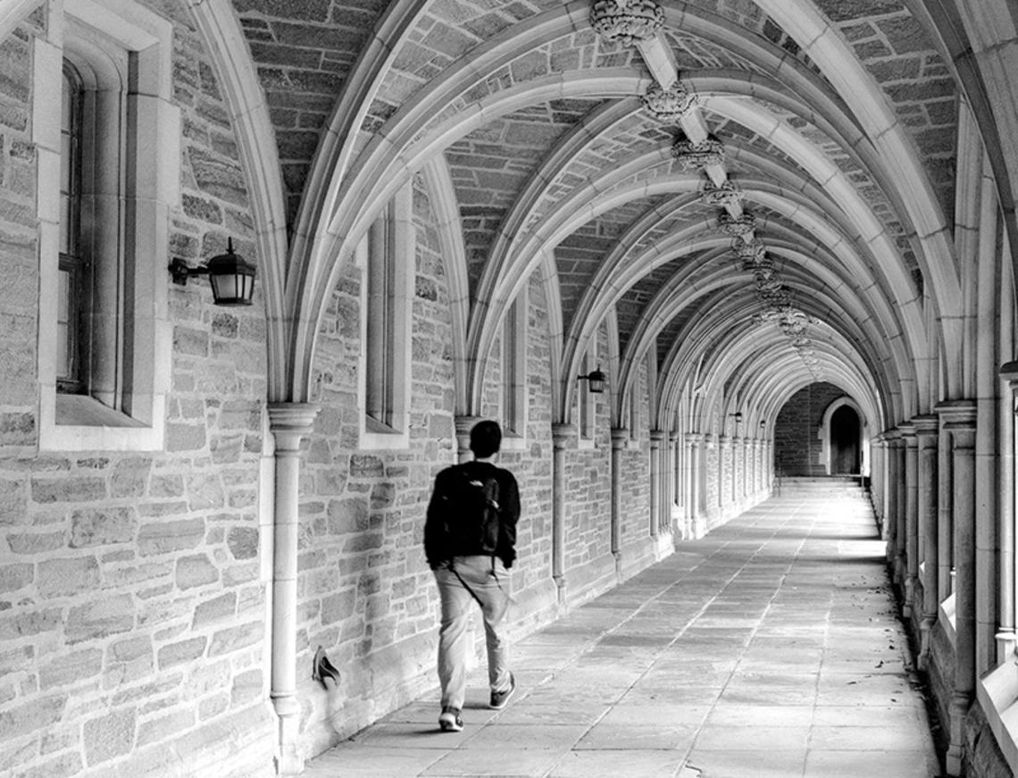 A black-and-white photo of a student walking down an arched stone hallway alone.