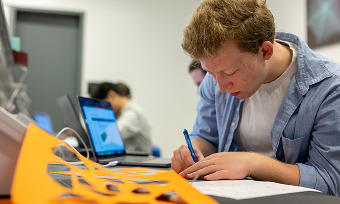 A student writes with a pen; a yellow piece of paper with shapes punched out is in the foreground.