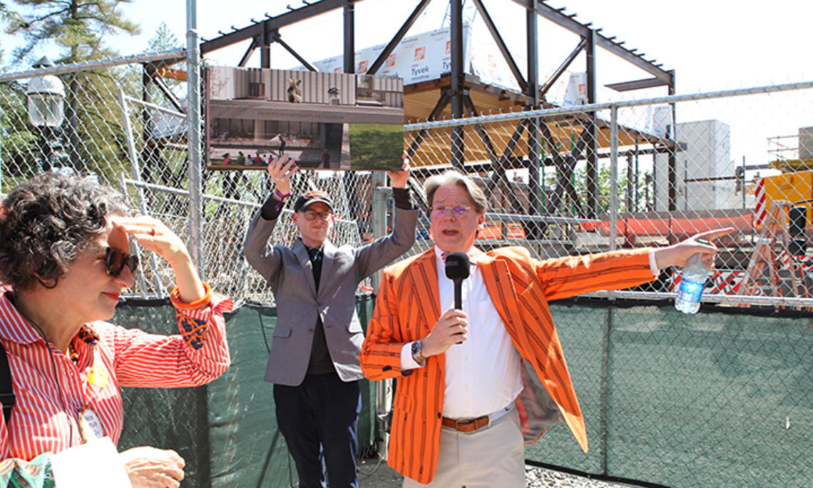 A man in an orange striped blazer speaks into a microphone in front of a construction site.
