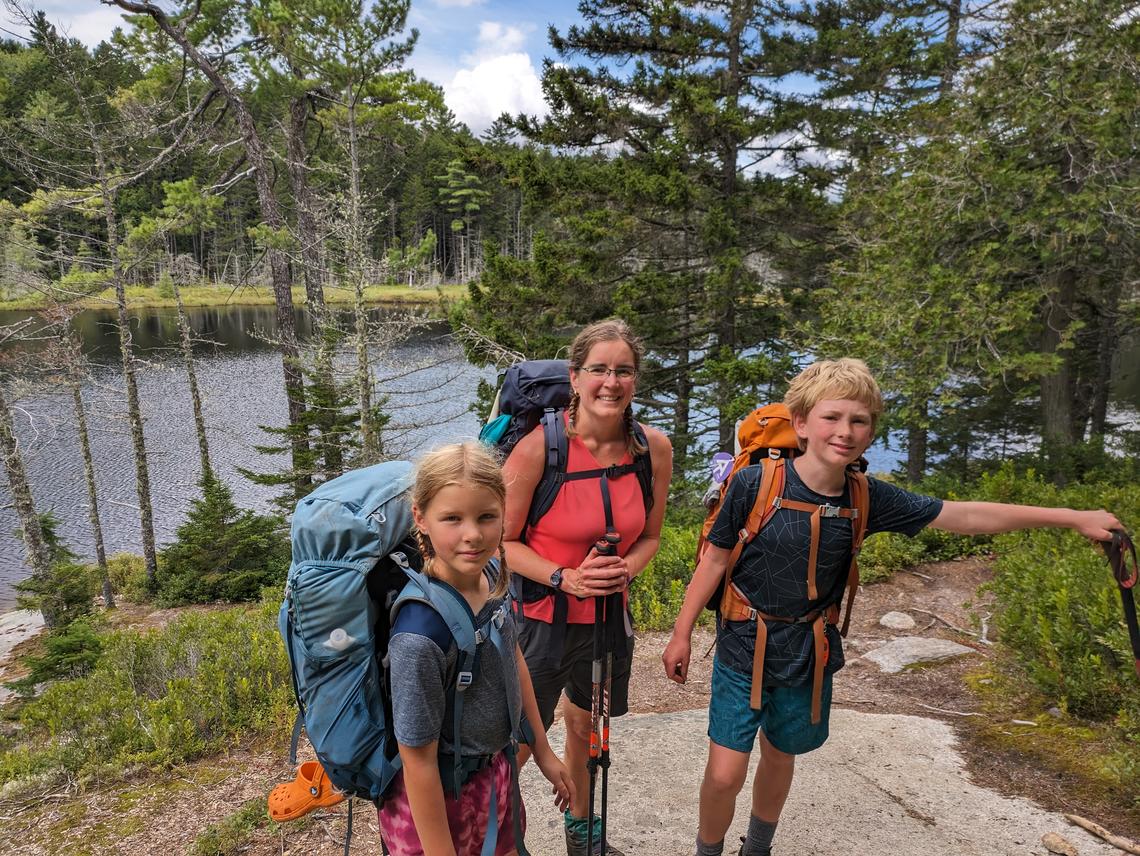 A woman and her two children wear backpacks on a trail; trees and a body of water are in the background.