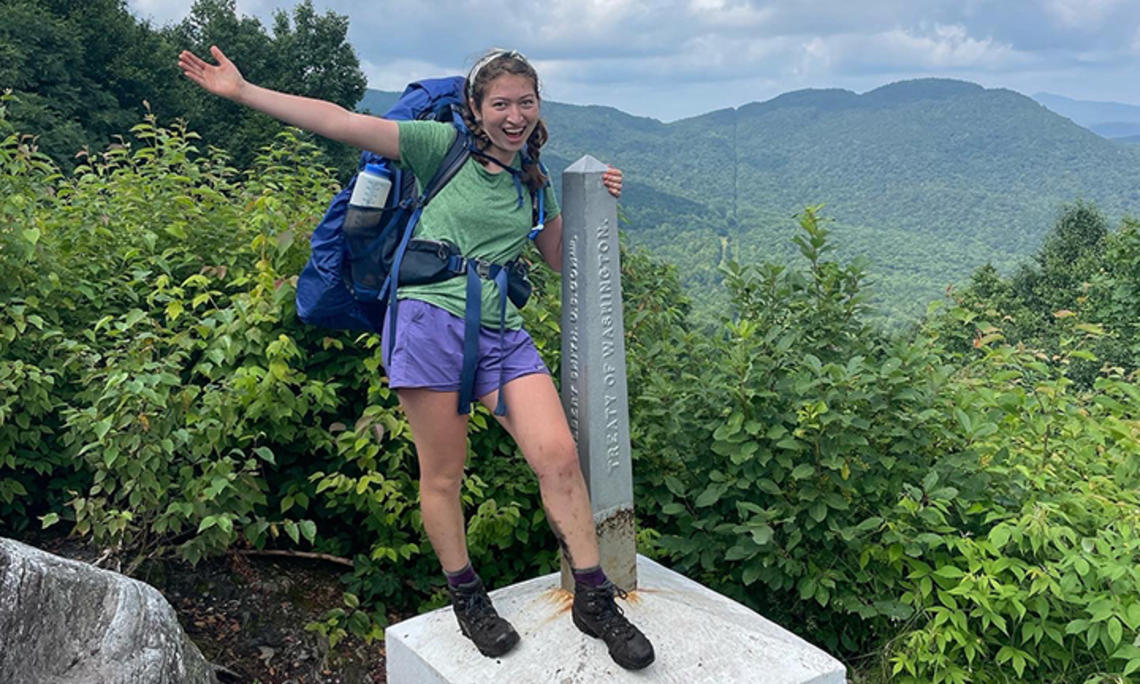 Wearing her backpack and hiking boots, Ruby Platt flings out an arm and smiles as she stands in front of the Green Mountains, holding onto an obelisk-shaped marker that reads "Treaty of Washington."