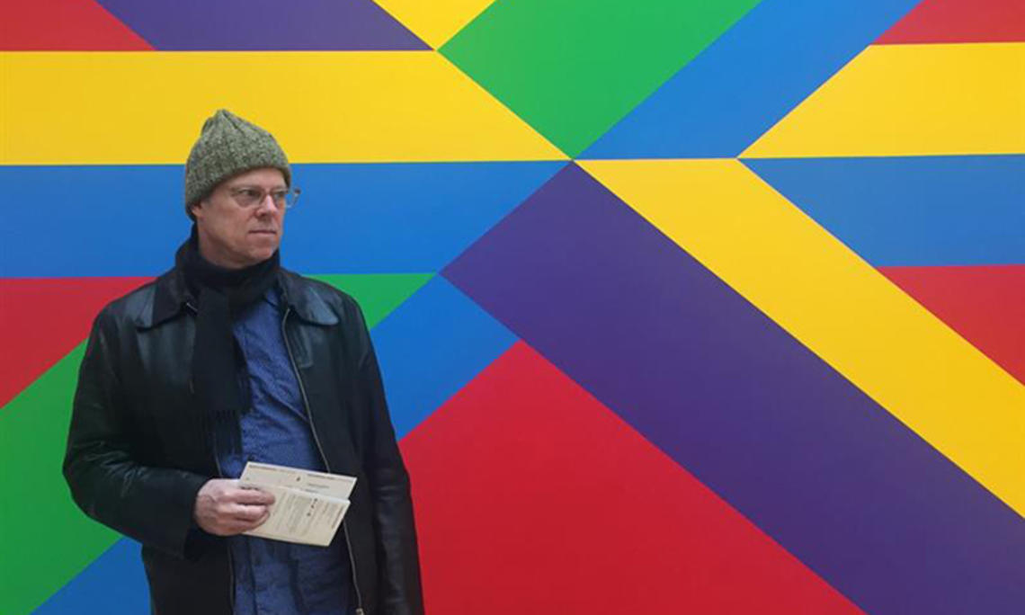 This is a photo of Joe Scanlan holding papers and standing in front of a brightly-painted wall.