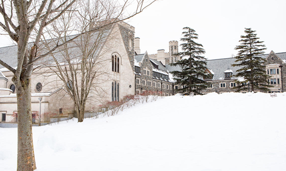 A view of part of Princeton’s Gothic stone campus, viewed up a snowy hill, with two large pine trees at the top.