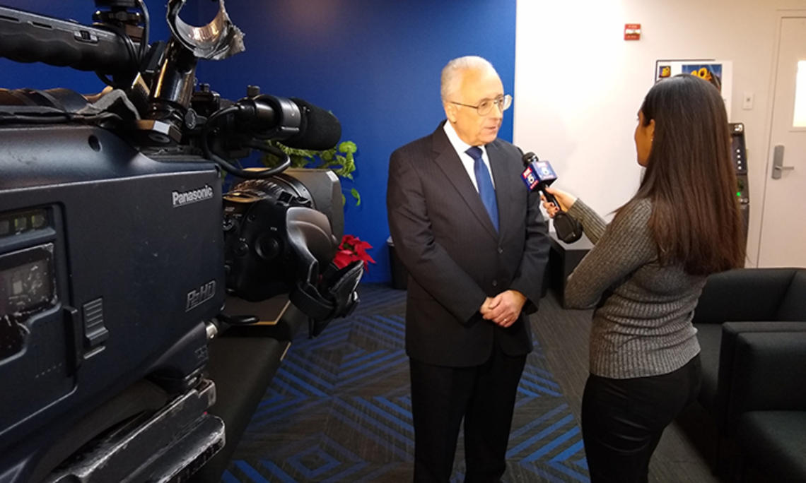 This is a photo of Alain Sanders being interviewed by a news reporter holding a microphone. A video camera filming them is in the foreground..