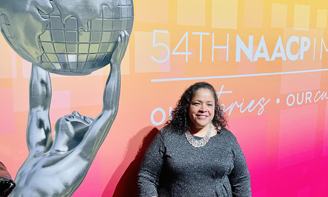 Janice Johnston ’92 stands in front of a large pink-and-yellow sign reading "54th NAACP." Part of the text is cut off. There's also a photo of a silver statue of Atlas holding up the globe.