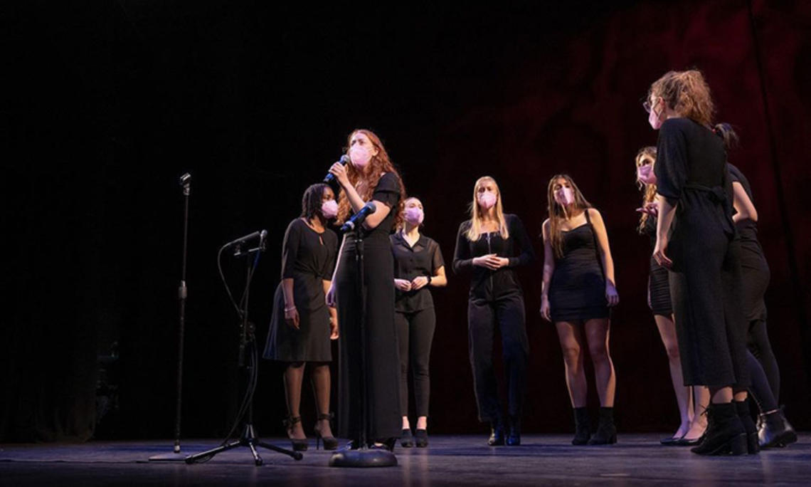 This is a photo of the Tigerlilies singing on stage; eight women are wearing black clothes and pink face masks while they sing.