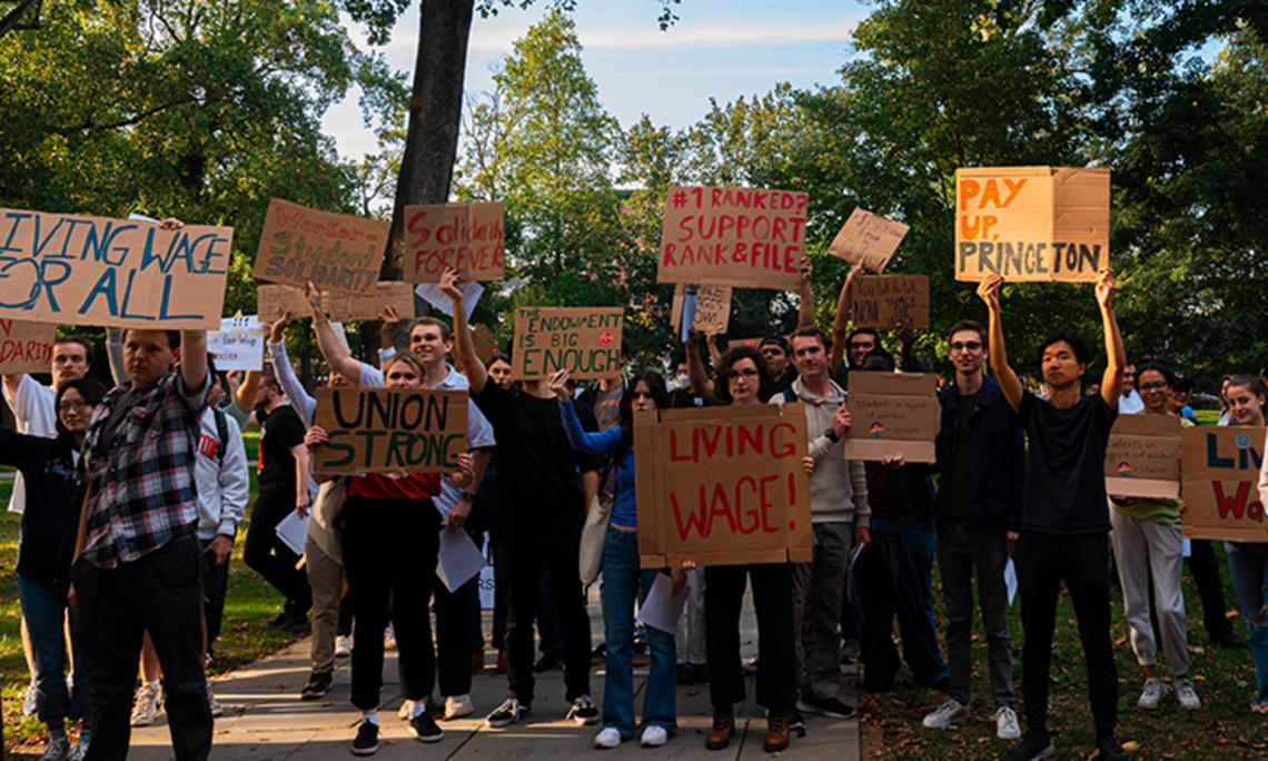 A crowd standing outside holds signs reading "Living Wage!" and "Pay Up Princeton" and #1 Ranked? Support Rank & File" and "Union Strong."