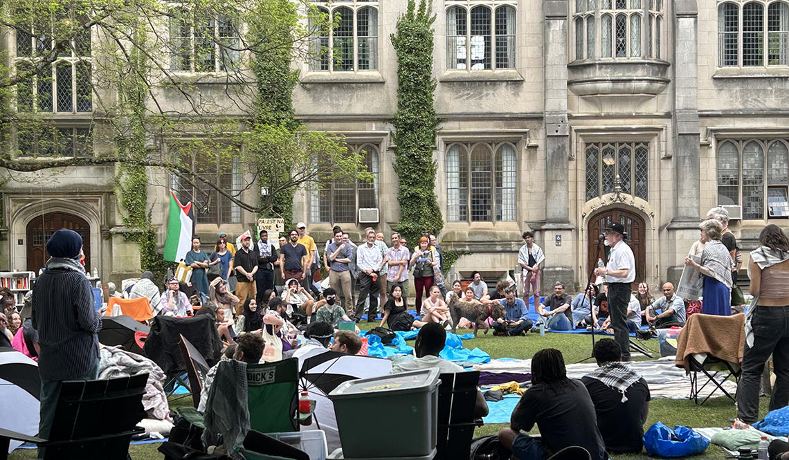 alum-0429.jpg A man stands at a microphone surrounded by protesters; a Gothic building is in the background.