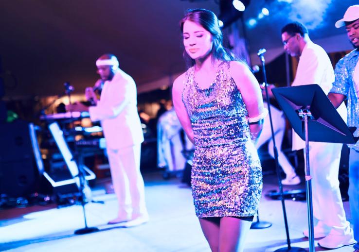 A woman on stage in a sparkly dress; men appear to play music in the background.