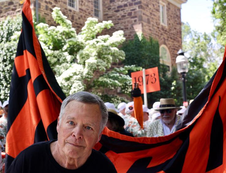 A man peers at the camera near an orange-and-black banner; someone in the background holds a sign reading "1973."